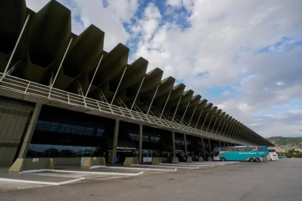 Exterior da Rodoviária Rita Maria em Florianópolis, SC, com cobertura em zigue-zague e ônibus intermunicipais estacionados sob céu nublado.