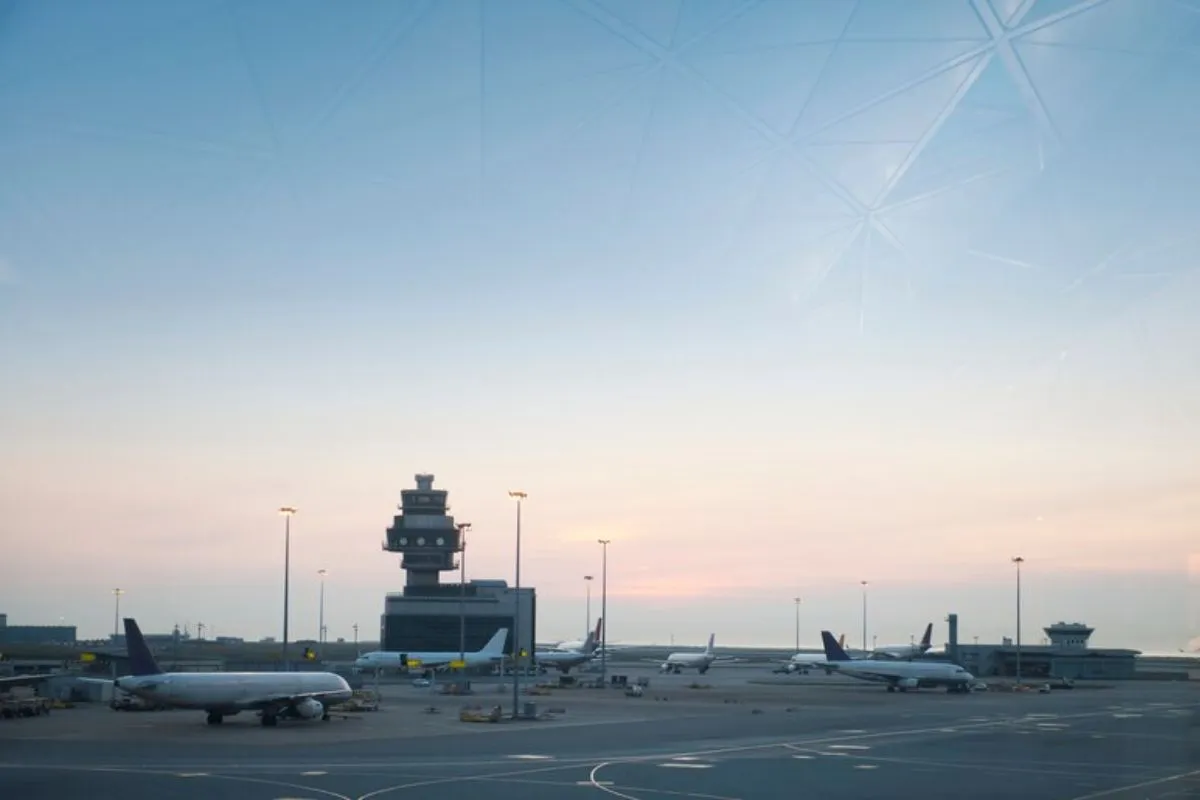 Aeroporto em imagem captada de longe com aeronaves pousadas do tipo bimotor à jato com torre de comando ao fundo da imagem em período de céu em penumbra.