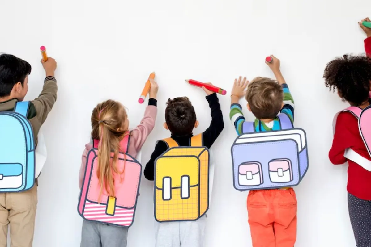 Cinco crianças de etnias, alturas e sexo diferentes de costas, portando mochilas semelhantes a desenhos gráficos enquanto seguram lápis de cores diversas e tamanho grande como se fossem desenhar na parede branca que está logo à frente deles.