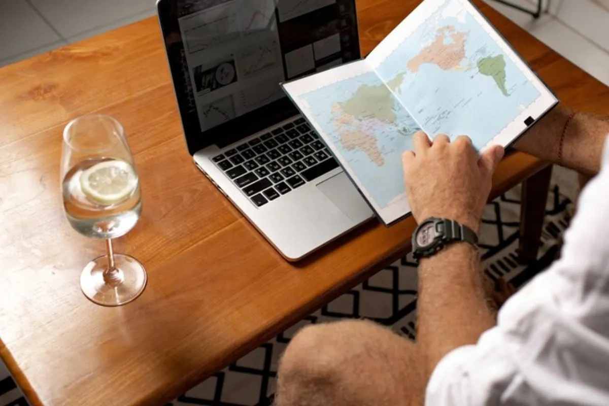 Homem sentado diante de mesa de madeira com notebook sobre a mesma e taça de vidro com água à sua esquerda, enquanto folheia um mapa em uma agenda.
