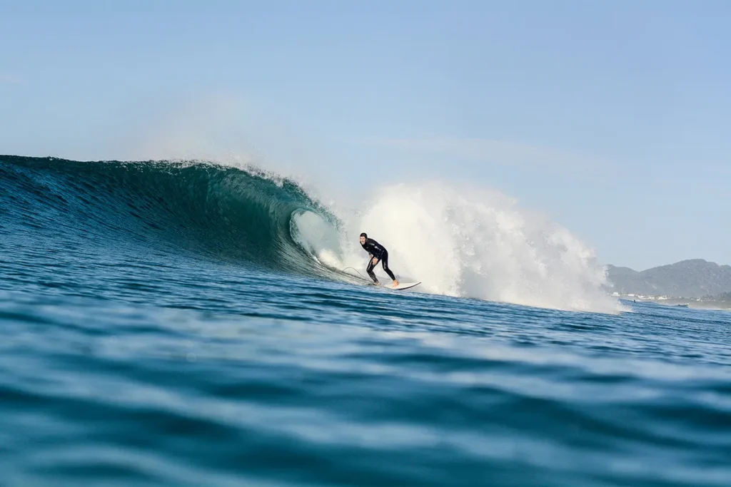 Um surfista, vestido com um traje de neoprene preto, está em pé em uma prancha branca, surfando uma grande onda azul e em formação na Praia da Joaquina. Uma paisagem montanhosa e casas são visíveis ao fundo, sob um céu azul e claro.