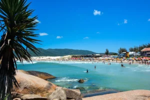 Uma vista da praia da Joaquina, com uma grande faixa de areia lotada de pessoas e barracas, com mar azul-turquesa e ondas. Ao fundo, dunas de areia branca e uma colina coberta de vegetação.
