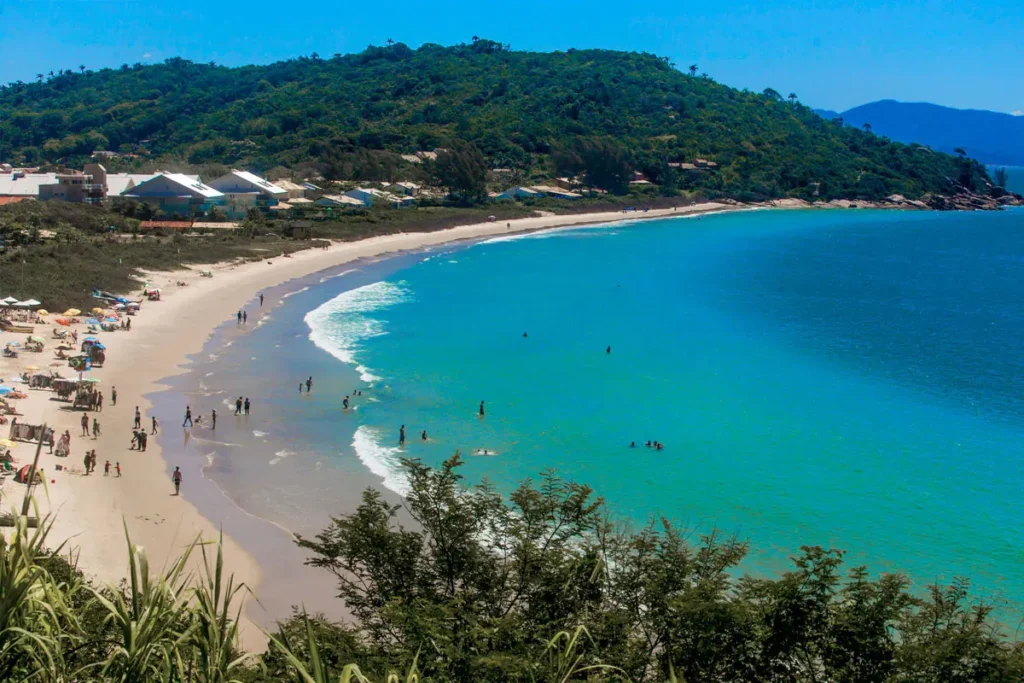 Vista panorâmica da praia da Lagoinha do Norte em Florianópolis, SC, com mar de águas cristalinas, faixa de areia populosa, vegetação costeira e colinas ao fundo