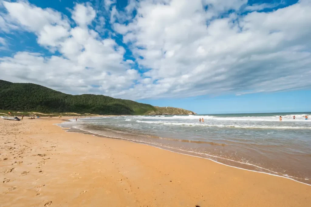 Praia da Lagoinha do Norte em Florianópolis, SC, com areia dourada, mar agitado, banhistas e montanhas cobertas por vegetação densa sob um céu com nuvens brancas.