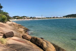 Vista da praia da Lagoinha do Norte em Florianópolis, SC, com pedras em primeiro plano, águas claras, faixa de areia com banhistas e a orla com casas e montanhas ao fundo.