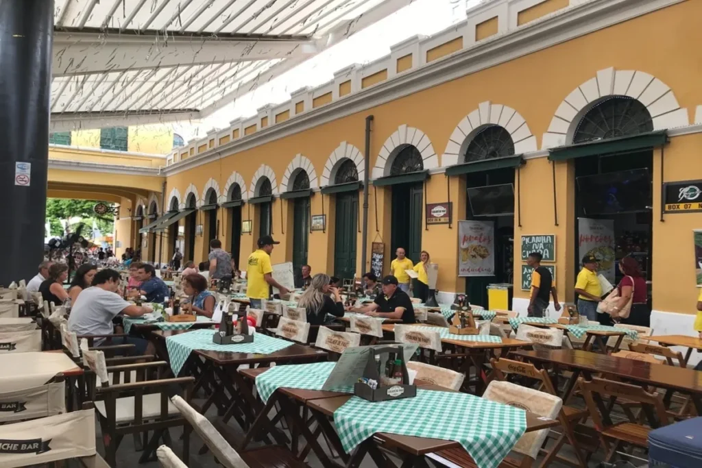 Área de alimentação do Mercado Público de Florianópolis, com mesas cheias de pessoas e arquitetura histórica amarela. Cultura e gastronomia.