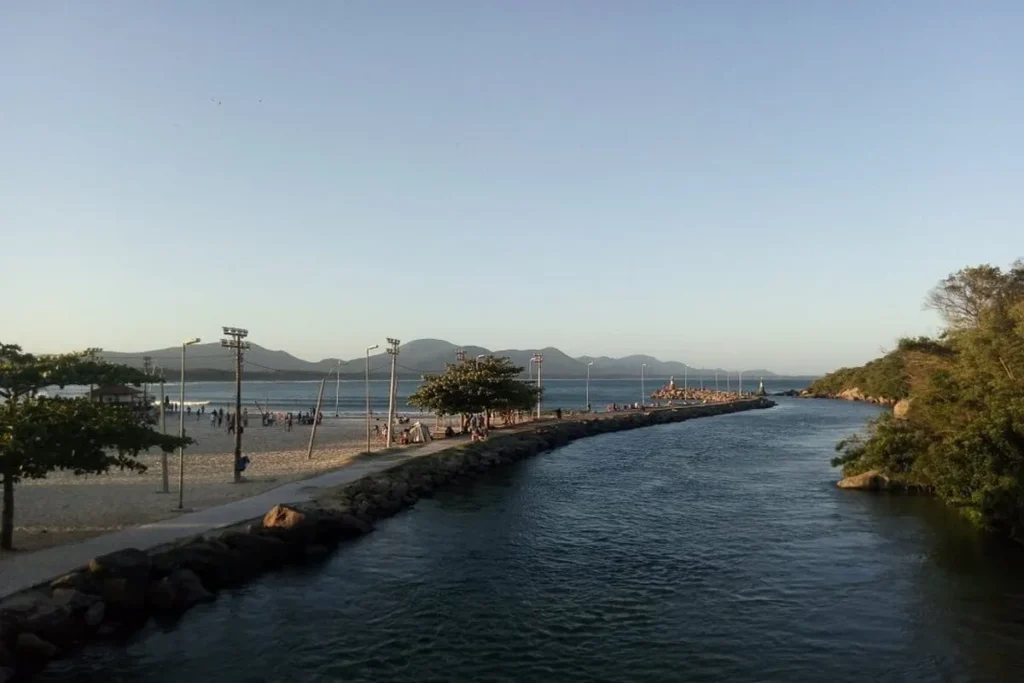 Vista do canal da Barra da Lagoa, em Florianópolis, com o mar ao fundo, um molhe rochoso com pessoas e um canal de águas calmas.