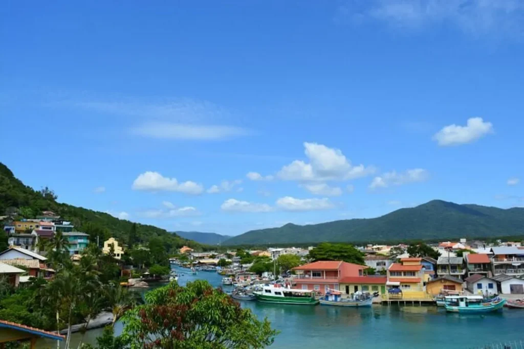 Vista aérea de um canal que serpenteia por uma paisagem urbana e montanhosa. Barcos de pesca estão atracados na margem, e casas de cores vibrantes se alinham na orla da Barra da Lagoa, em Florianópolis.
