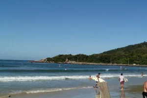 Vista da praia da Barra da Lagoa, em Florianópolis, com pessoas nadando e surfando. Dois surfistas, em primeiro plano, seguram suas pranchas, e um morro verde se estende ao fundo.