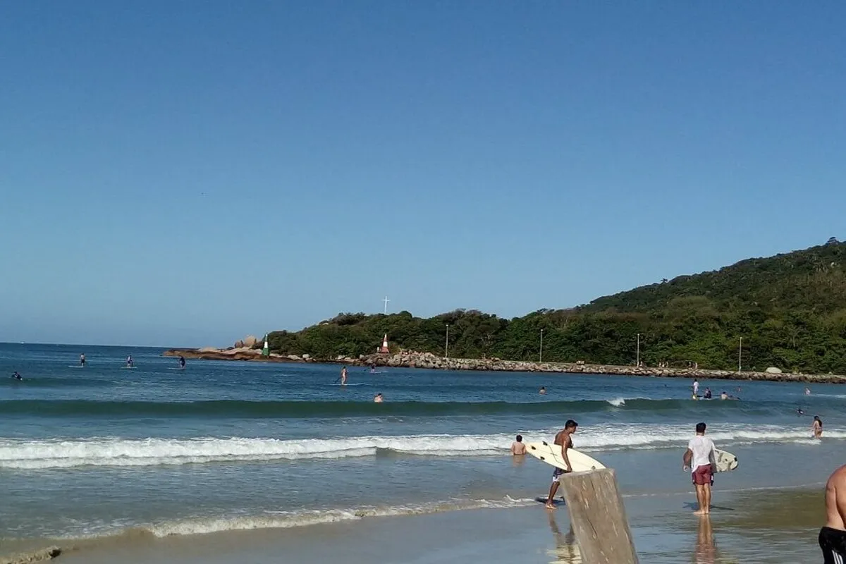 Vista da praia da Barra da Lagoa, em Florianópolis, com pessoas nadando e surfando. Dois surfistas, em primeiro plano, seguram suas pranchas, e um morro verde se estende ao fundo.