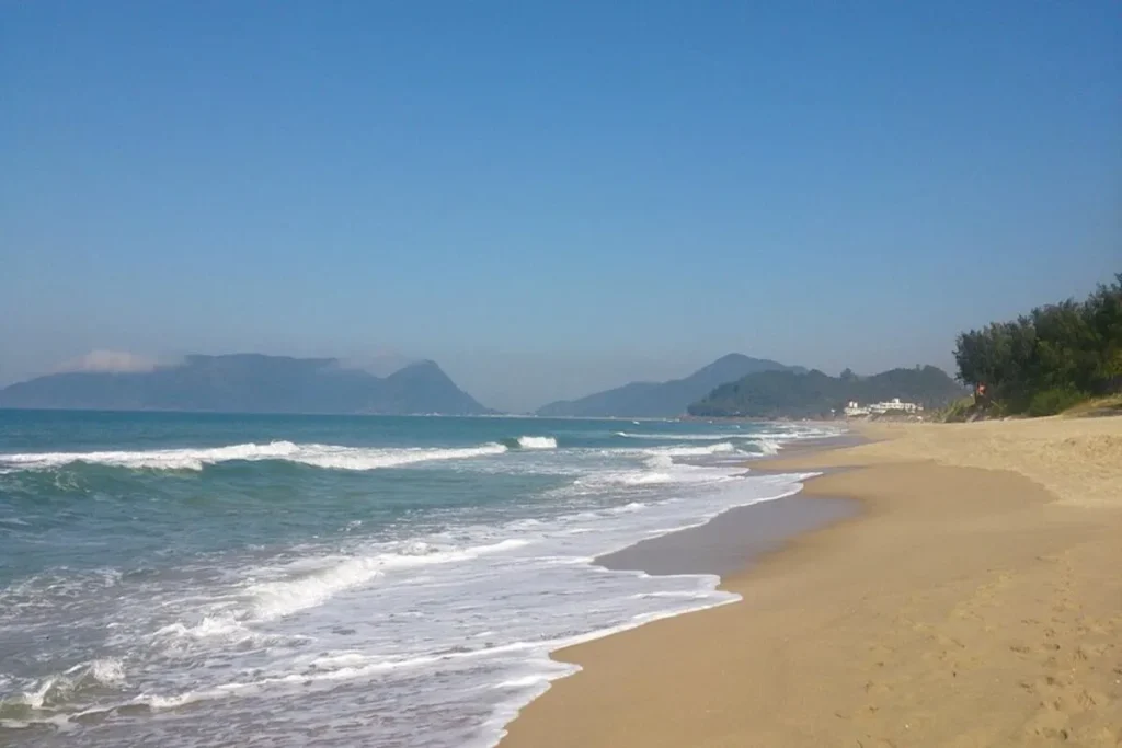 A praia do Campeche em Florianópolis, com areia dourada e mar calmo, com morros ao fundo.