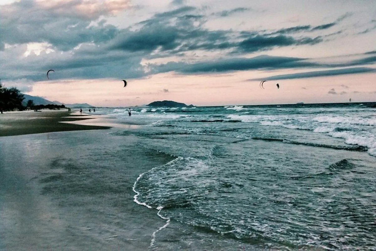A praia do Campeche em Florianópolis, com o mar calmo e a prática de kitesurfe ao fundo.