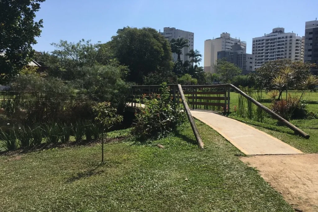 Caminho com uma pequena ponte de madeira em um Jardim Botânico, com prédios e arranha-céus visíveis ao fundo, mostrando a natureza em ambiente urbano.