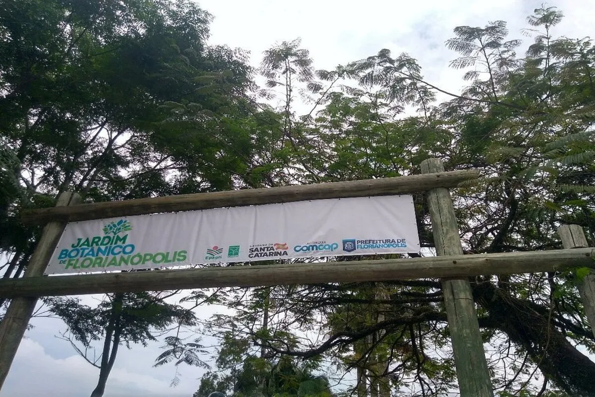 Placa de entrada do Jardim Botânico de Florianópolis feita de madeira rústica, com o nome do local e logotipos institucionais, indicando o acesso ao parque.