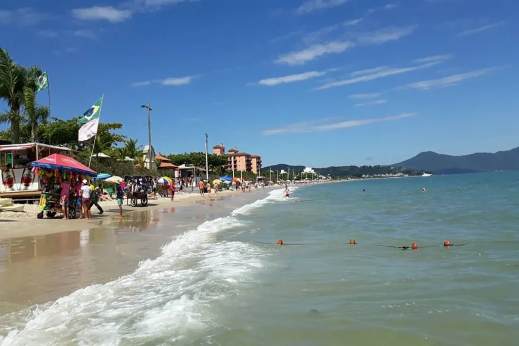 Jurerê, praia com areia branca, mar azul-claro, guarda-sóis coloridos e pessoas relaxando.