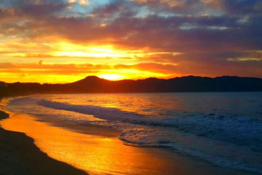 Jurerê, pôr do sol dourado em uma praia com ondas e silhueta de montanhas.