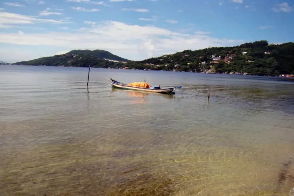 Vista da Lagoa da Conceição em Florianópolis, com um barco de pesca tradicional ancorado em águas claras e a paisagem de montanhas ao fundo.