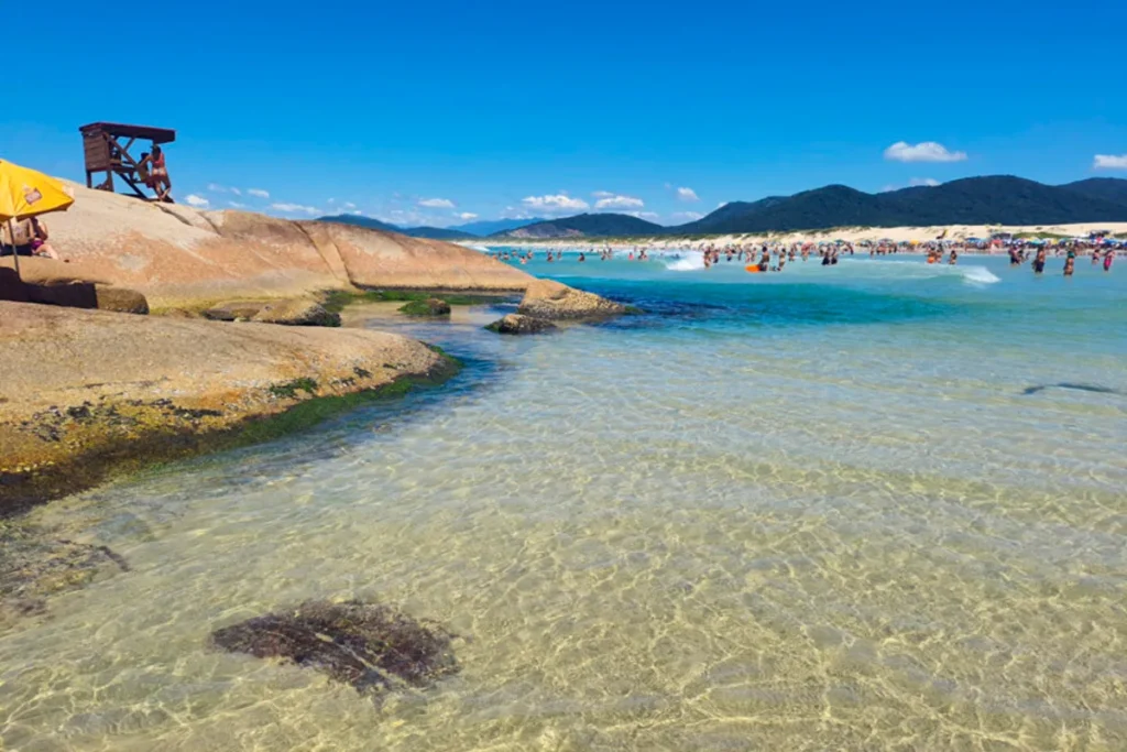 Vista da Lagoa da Conceição, com uma praia de areia lotada de pessoas, pedras grandes no canto esquerdo e a água transparente do mar.
