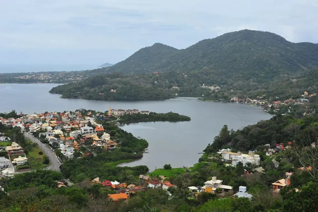 Vista panorâmica da Lagoa da Conceição em Florianópolis, com a lagoa, a cidade e as montanhas ao fundo, sob um céu nublado.
