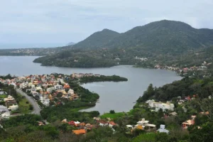 Vista panorâmica da Lagoa da Conceição em Florianópolis, com a lagoa, a cidade e as montanhas ao fundo, sob um céu nublado.