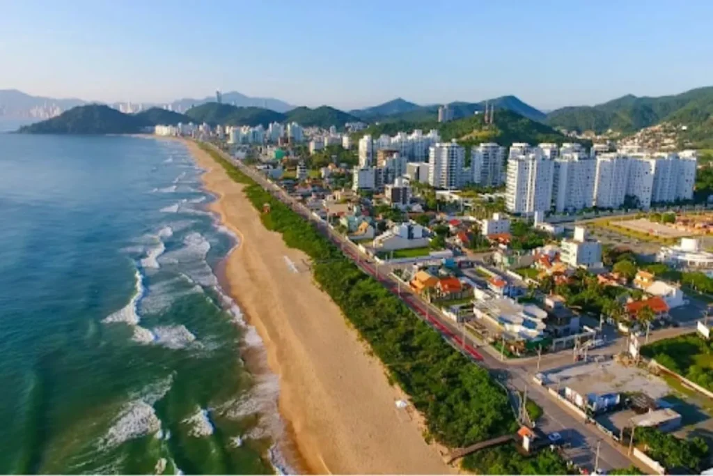 Vista aérea da Praia Brava, em Florianópolis, mostrando a longa orla de areia, a avenida costeira e os edifícios à beira-mar.