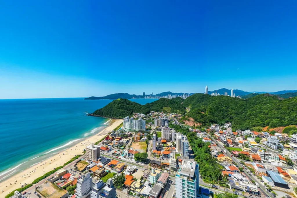 Vista aérea panorâmica da Praia Brava, com a longa faixa de areia, a avenida costeira e edifícios à beira-mar.