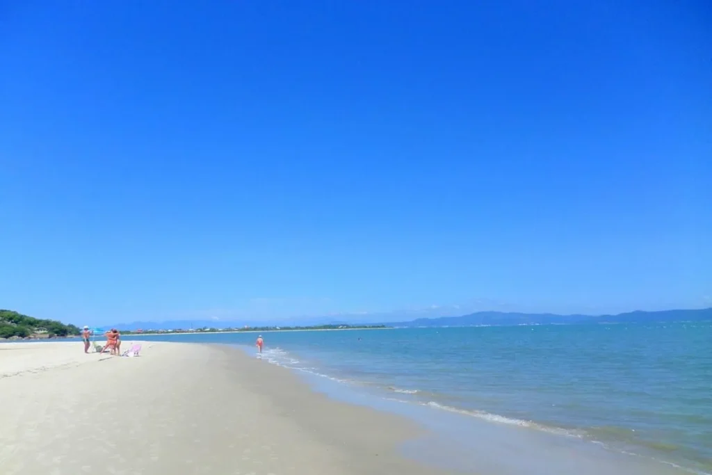 Uma vista panorâmica e serena da Praia do Forte em Florianópolis, Santa Catarina, em um dia ensolarado. A imagem mostra uma ampla faixa de areia clara e vazia na beira do mar calmo, com o horizonte pontuado por montanhas suaves. À esquerda, um pequeno grupo de pessoas aproveita a praia, enquanto uma única criança se diverte na água, reforçando a atmosfera de tranquilidade e relaxamento.
