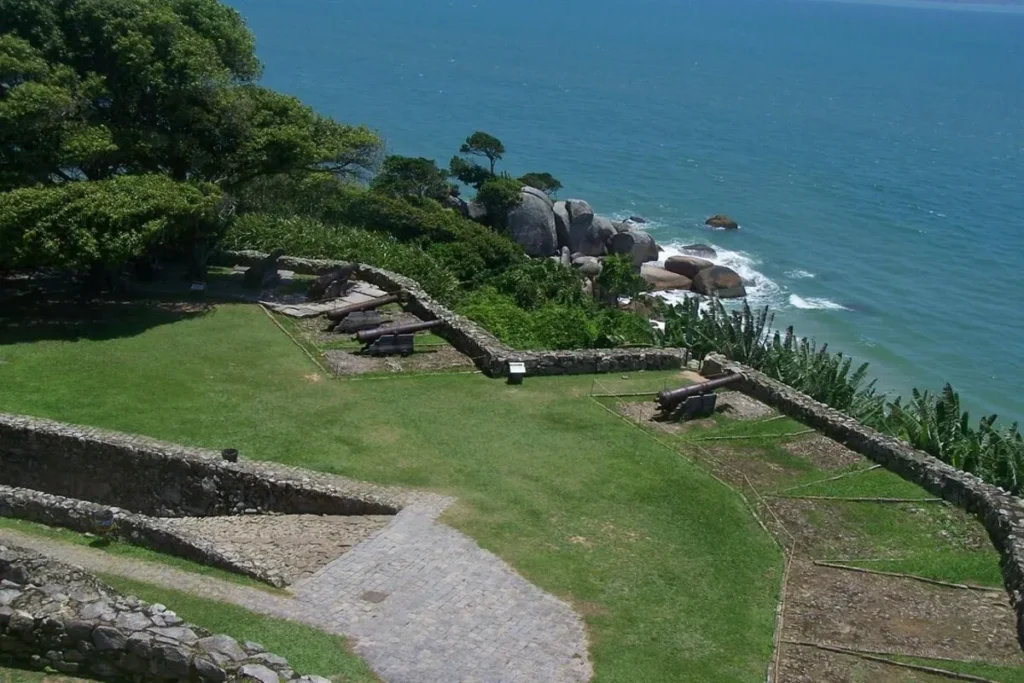 Uma vista aérea do Forte de São José da Ponta Grossa, próximo à Praia do Forte, em Florianópolis. A imagem destaca a estrutura histórica com seus canhões antigos apontando para o mar azul e rochoso. O gramado verde e bem cuidado do forte contrasta com a vegetação exuberante e as imponentes formações rochosas na encosta, que se encontram com as águas límpidas do oceano, criando uma cena que une história e beleza natural.