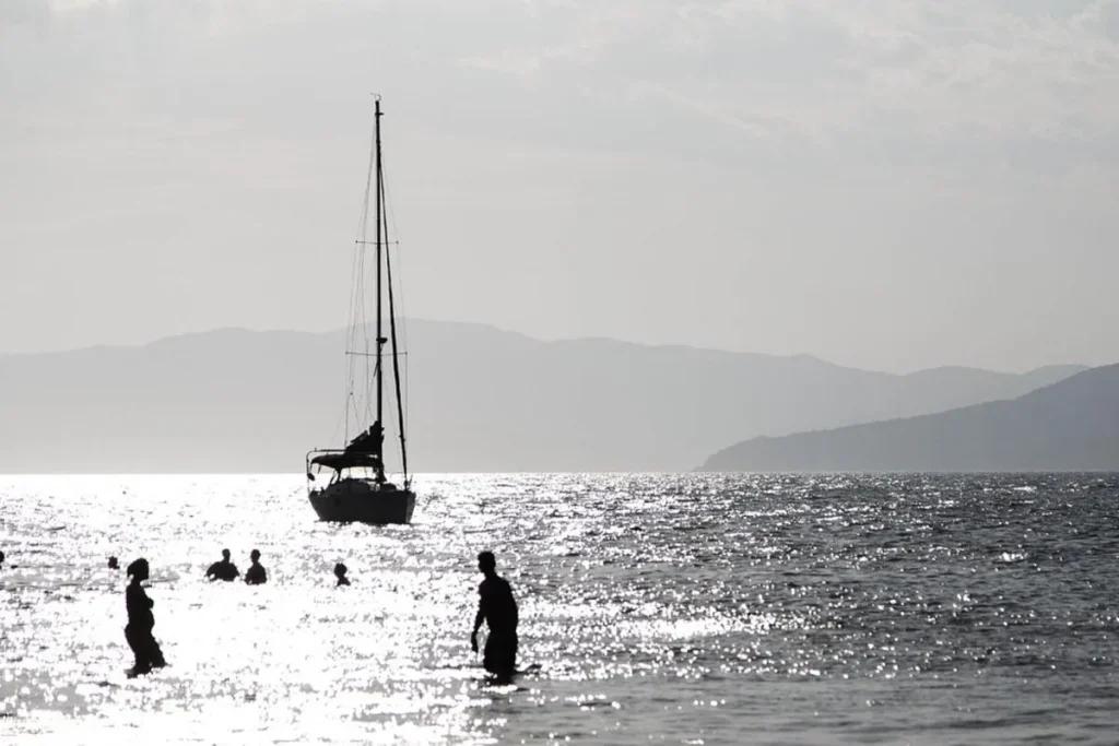 Uma imagem em contraluz da Praia do Forte em Florianópolis, Santa Catarina, capturando um veleiro ancorado no mar, com seu mastro alto se destacando contra o céu claro. O sol reflete intensamente na água, criando um brilho prateado que ilumina silhuetas de pessoas nadando e caminhando. Montanhas distantes adicionam profundidade ao horizonte, completando a cena tranquila e dramática.