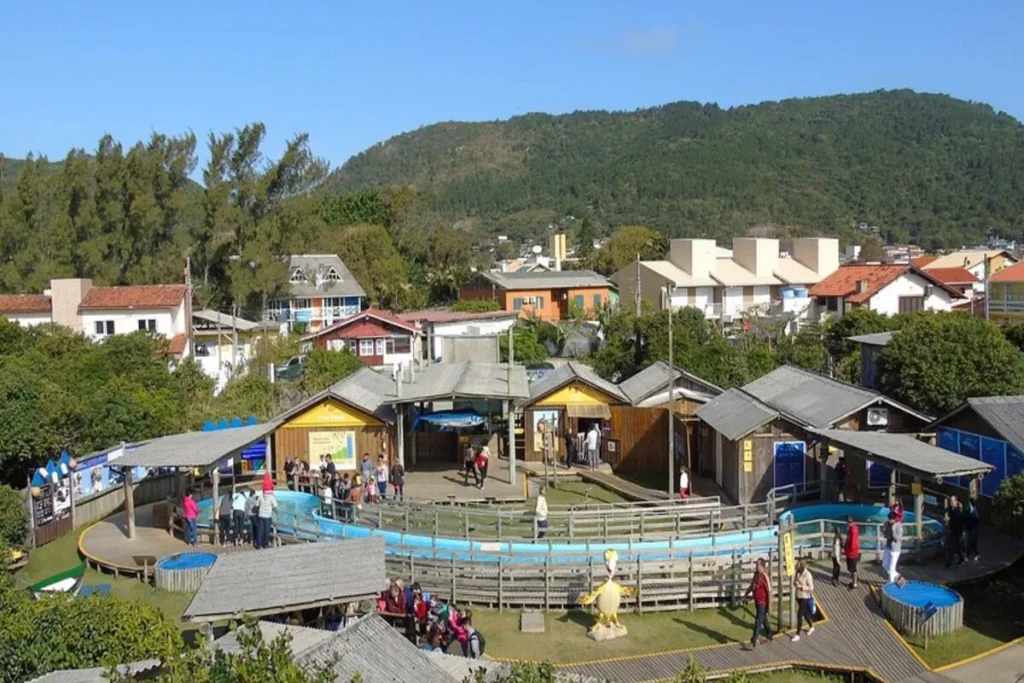 Vista aérea do centro de visitantes do Projeto Tamar, com tanques de água circulares, edifícios de madeira, pessoas e uma paisagem com casas e montanhas ao fundo.