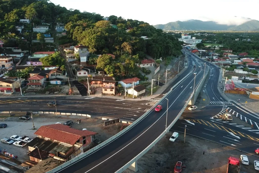 Vista aérea de um viaduto e complexo de estradas modernas no Rio Tavares, com casas em encostas e montanhas ao fundo.