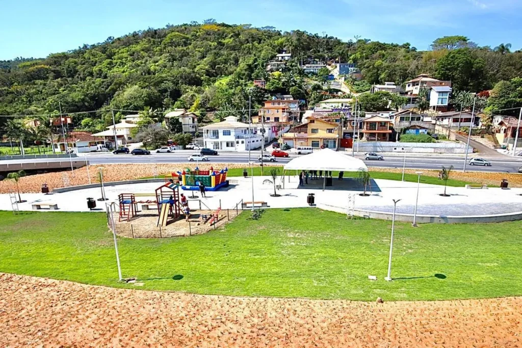 Praça pública e área de lazer com parquinho no Rio Tavares, com estrada movimentada e casas na encosta de um morro ao fundo.