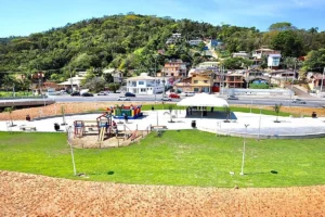 Praça pública e área de lazer com parquinho no Rio Tavares, com estrada movimentada e casas na encosta de um morro ao fundo.