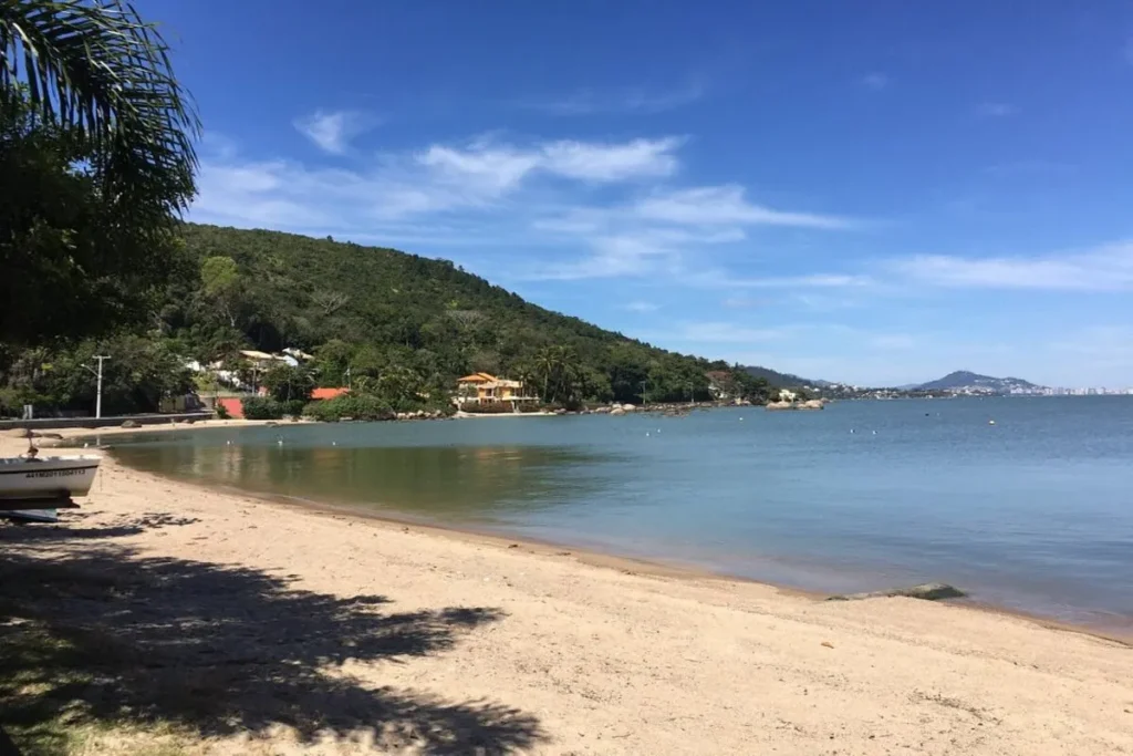 Praia de Sambaqui em um dia ensolarado, com areia clara, água calma e uma casa na encosta de um morro ao fundo.