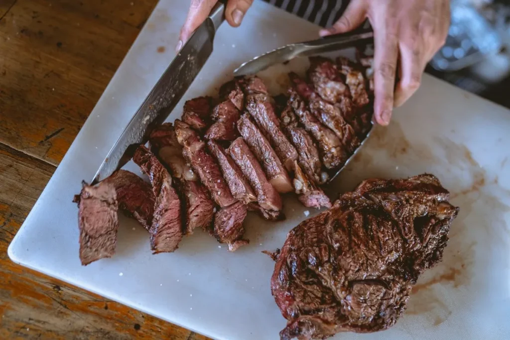 Mãos fatiando carne grelhada em uma tábua de corte branca.