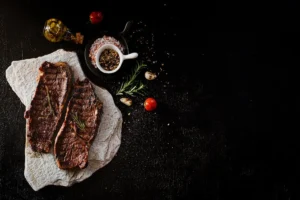 Dois bifes grelhados em uma pedra, com temperos, alho e tomate, em um fundo preto.