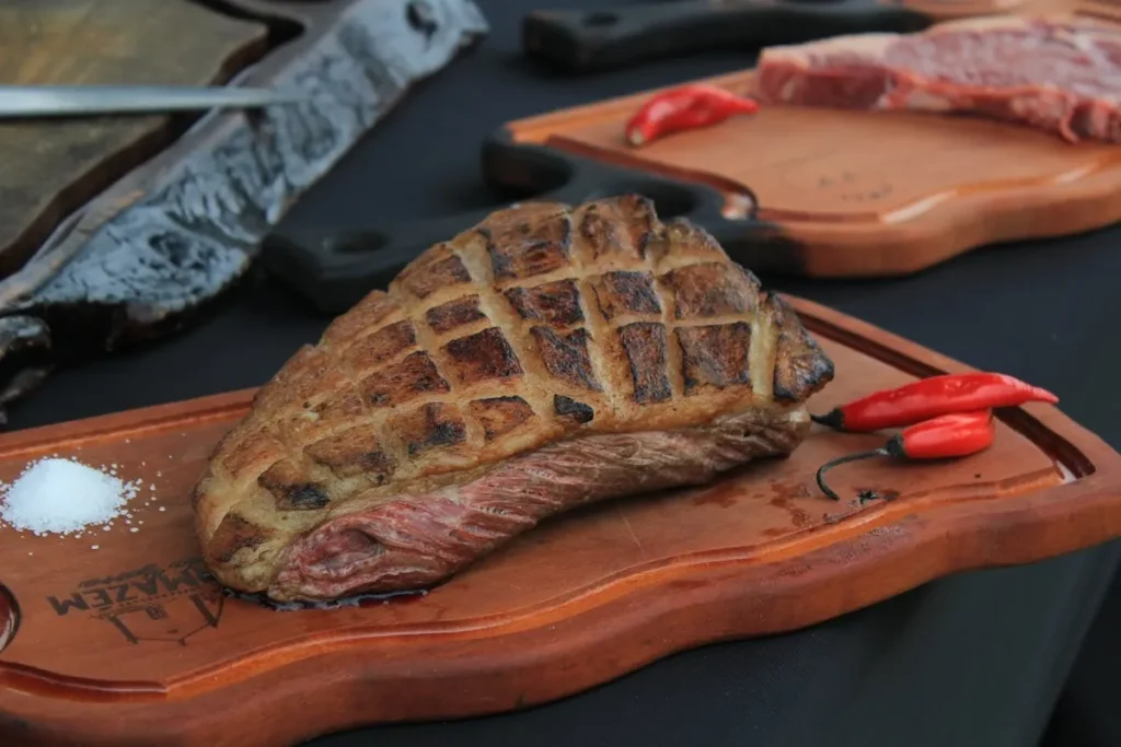 Corte de picanha grelhada com marcas de grelha em uma tábua de madeira com sal e pimenta vermelha, representando a culinária de churrascarias.