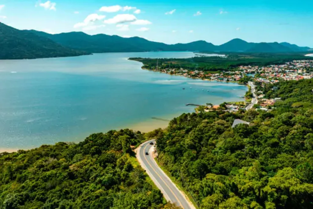 Vista aérea panorâmica da Lagoa da Conceição em Florianópolis, mostrando a lagoa, a praia, a cidade e as montanhas ao fundo.