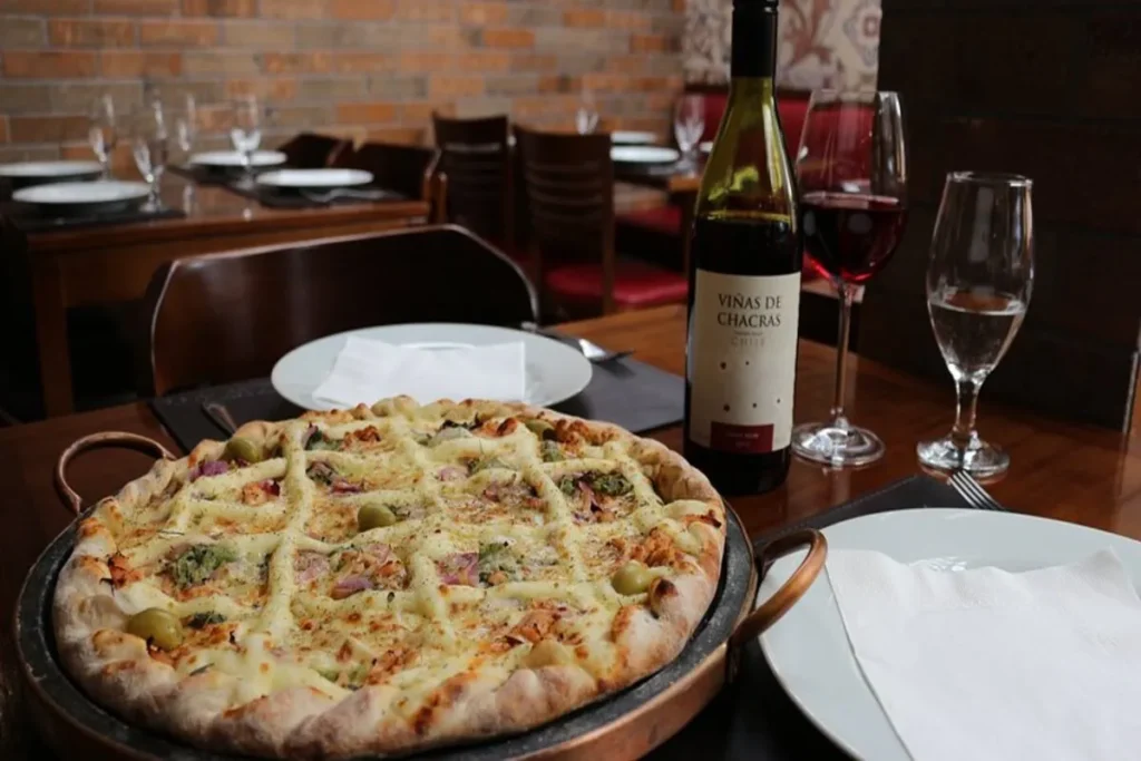Uma pizza com cobertura elaborada, servida em uma travessa de cobre, sobre uma mesa de restaurante com uma garrafa de vinho tinto e taças. A imagem representa uma opção de pizza gourmet em Florianópolis.
