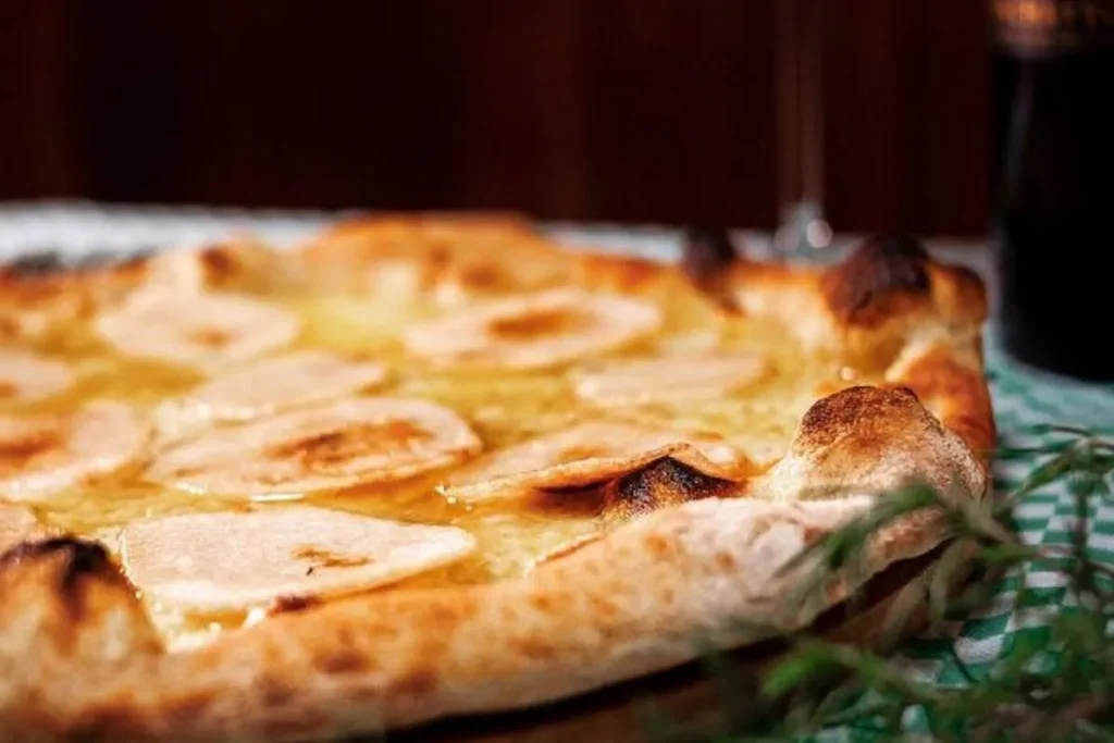 Uma pizza de sobremesa com fatias de pera ou maçã e uma cobertura brilhante, possivelmente mel ou queijo, servida em uma mesa de restaurante com um copo de vinho ao fundo. A imagem representa uma pizza doce gourmet em Florianópolis.