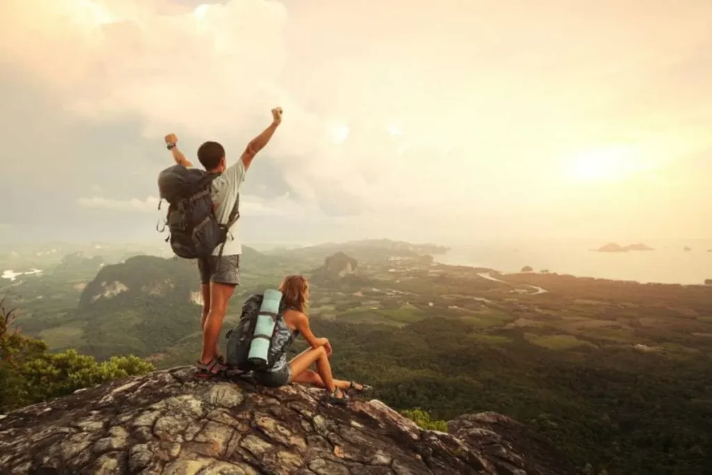 Homem comemorando no topo de um rochedo e mulher sentada, ambos de mochila, observando a vasta paisagem verde e o oceano durante o pôr do sol.