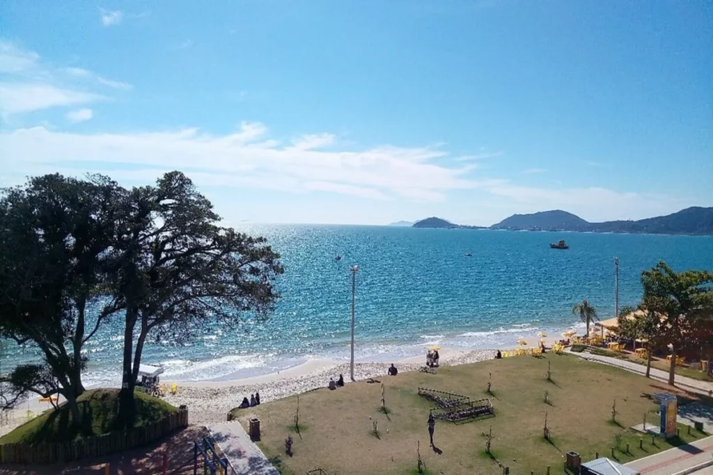 A fotografia retrata o pôr do sol na Praia de Canasvieiras, em Florianópolis, Santa Catarina. O sol se despede no horizonte, refletindo sua luz dourada sobre o mar calmo. Em primeiro plano, surgem silhuetas de árvores, quiosques e a faixa de areia, enquanto montanhas ao fundo completam o cenário paradisíaco que transmite paz e beleza natural.