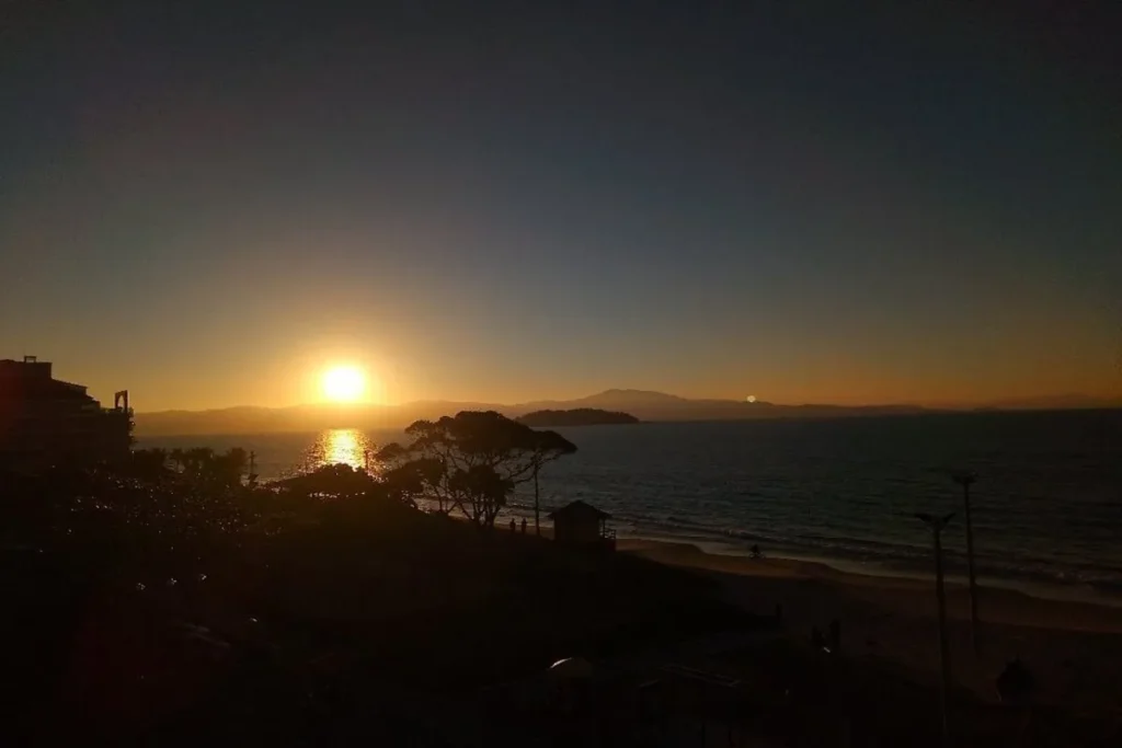 Pôr do sol na Praia de Canasvieiras, em Florianópolis, com vista do mar, árvores e montanhas ao fundo.