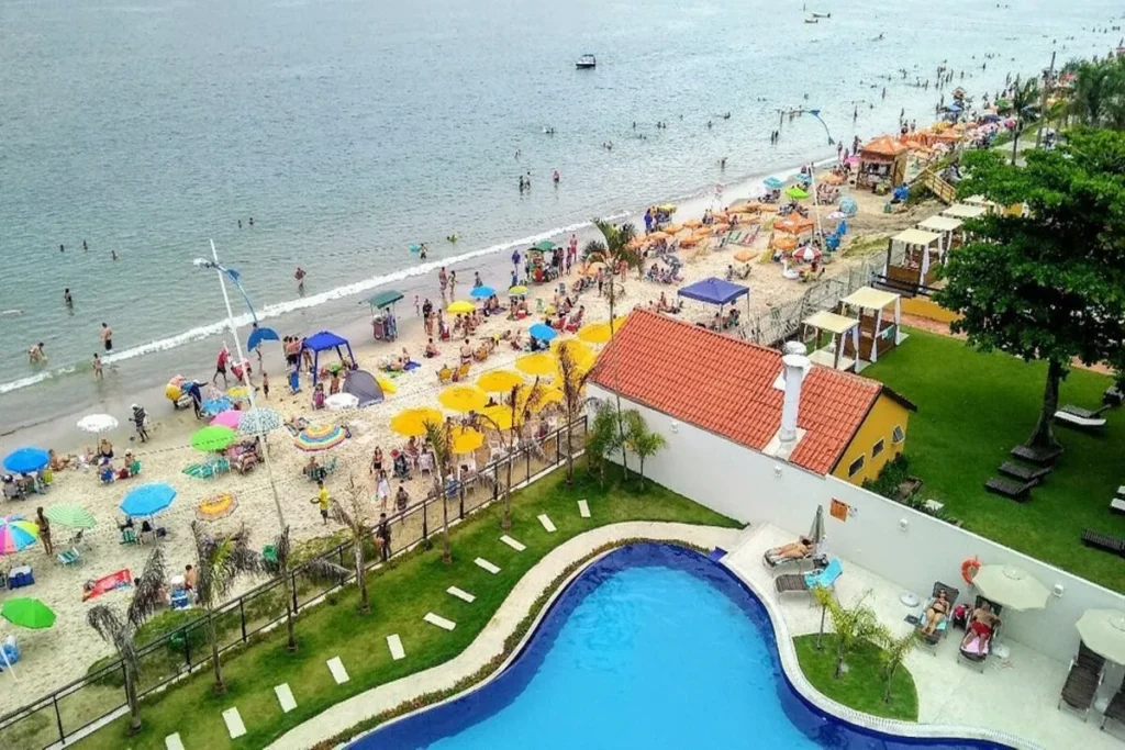 Praia de Canasvieiras em Florianópolis com guarda-sóis coloridos, banhistas no mar e piscina de hotel em frente à orla.