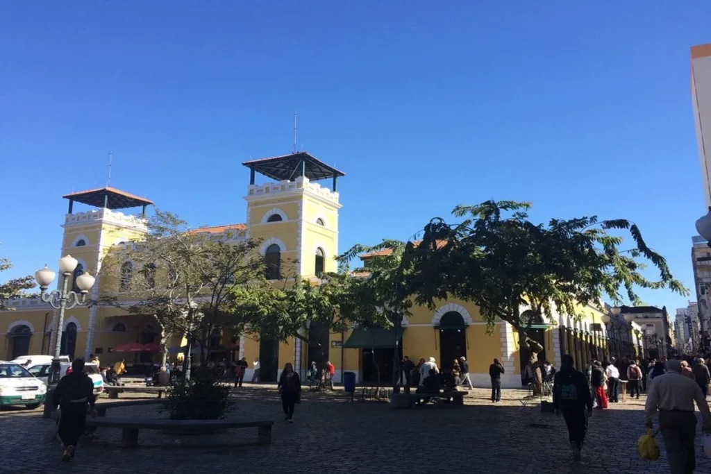 Mercado Público de Florianópolis, um prédio histórico de cor amarela, com duas torres, em frente a uma praça com árvores e pessoas.