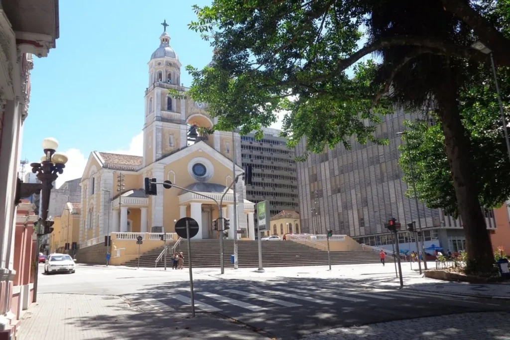Catedral Metropolitana de Florianópolis, de cor amarela, em frente a uma praça com escadaria e um prédio moderno ao lado, sob um céu azul.
