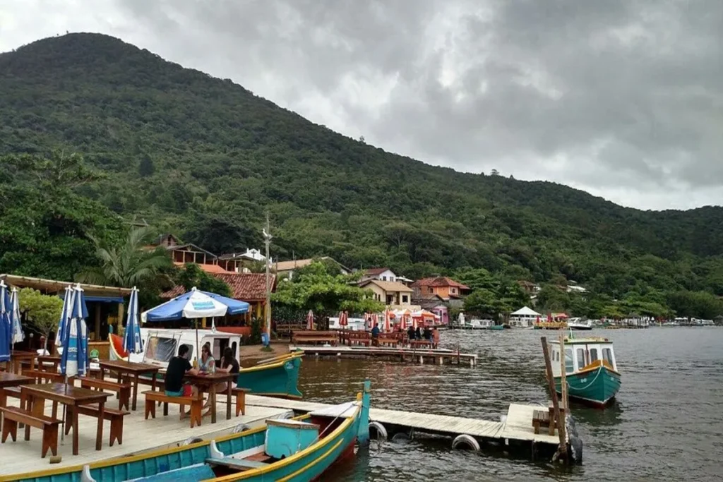 Píer rústico com barcos coloridos e mesas de restaurante à beira-mar na Costa da Lagoa, em Florianópolis, com um morro coberto de mata ao fundo.