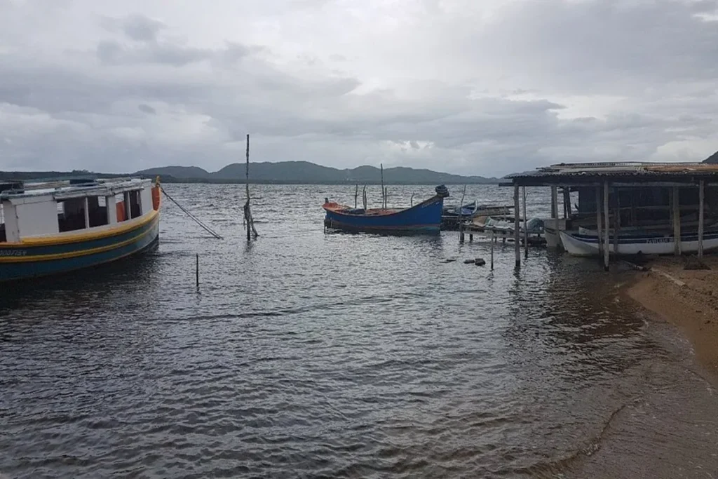 Vista de um píer e barcos rústicos na Costa da Lagoa, em Florianópolis, com a lagoa e morros ao fundo sob um céu nublado.