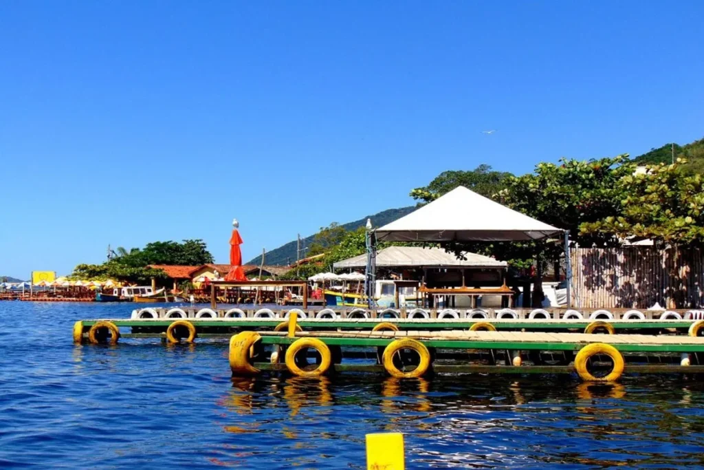 Píer rústico de madeira com boias de pneus, barcos e restaurantes à beira da Costa da Lagoa em Florianópolis, sob um céu azul.