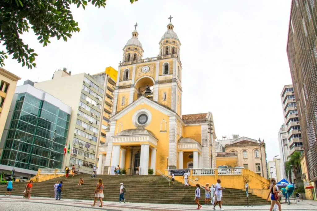 A Catedral Metropolitana de Florianópolis, um edifício religioso de cor amarela e branca, vista de uma praça movimentada. O prédio tem duas torres sineiras altas e uma grande escadaria frontal. Várias pessoas estão subindo e descendo os degraus e andando na praça. Ao redor da catedral, há prédios modernos e altos. O céu está claro e o dia parece nublado.